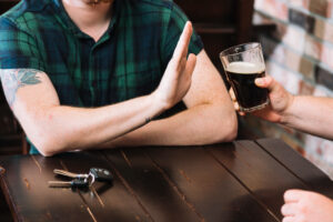 Person making a stop gesture to refuse an offered glass of dark beer, with car keys visible on table symbolizing responsible decision-making