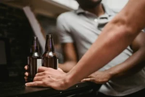 A close-up of a person's hand resting on a table next to two brown glass bottles, with another person blurred in the background.